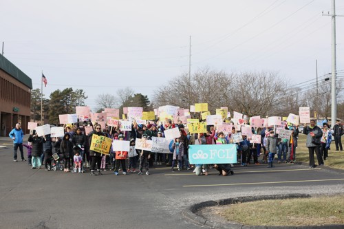 A crowd of people from the 2023 M.L.K. Legacy March, holding signs outside. In the center is a sign with the  F.C.L logo